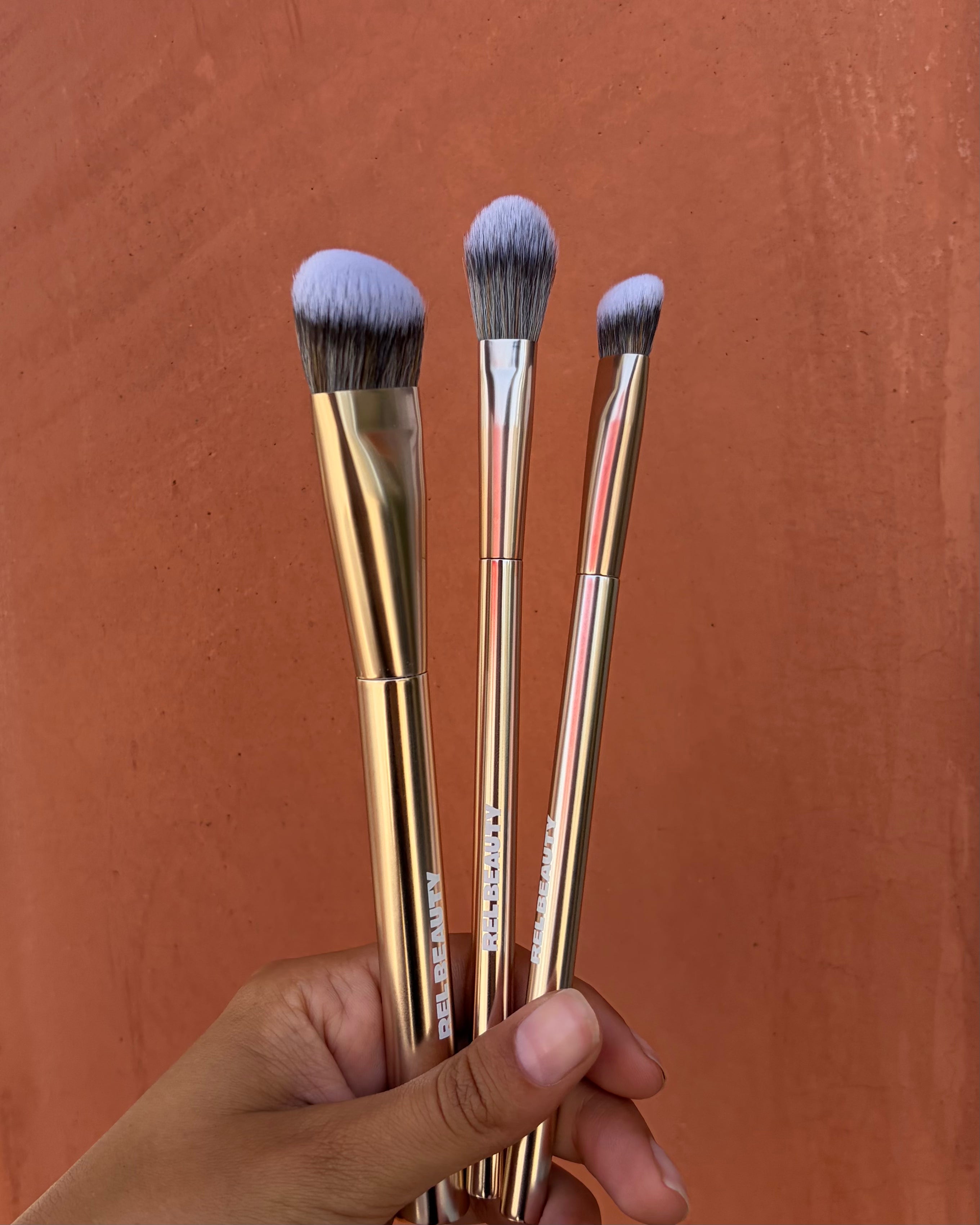 Three makeup brushes with gold handles and gray-purple bristles, held in a hand against a textured terracotta background.