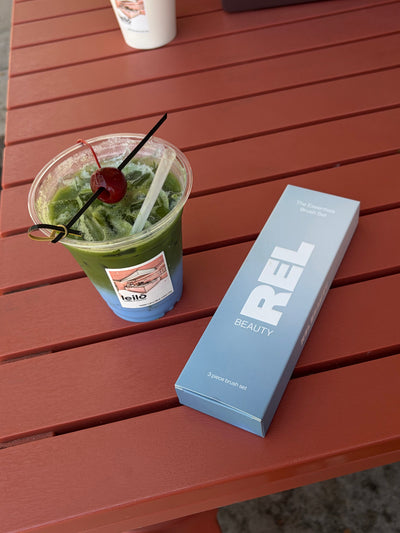 A light blue REL BEAUTY product box for 'The Essentials Brush Set' sits on a reddish-brown slatted table next to a green drink with a cherry garnish.
