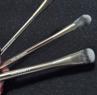 Three makeup brushes with metallic gold handles and black bristles, branded REL BEAUTY, resting on a dark textured background.